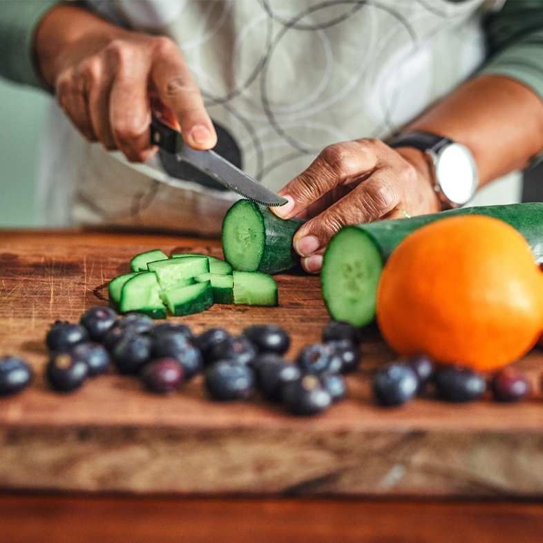 Person cutting cucumber with an orange and blueberries also on cutting board