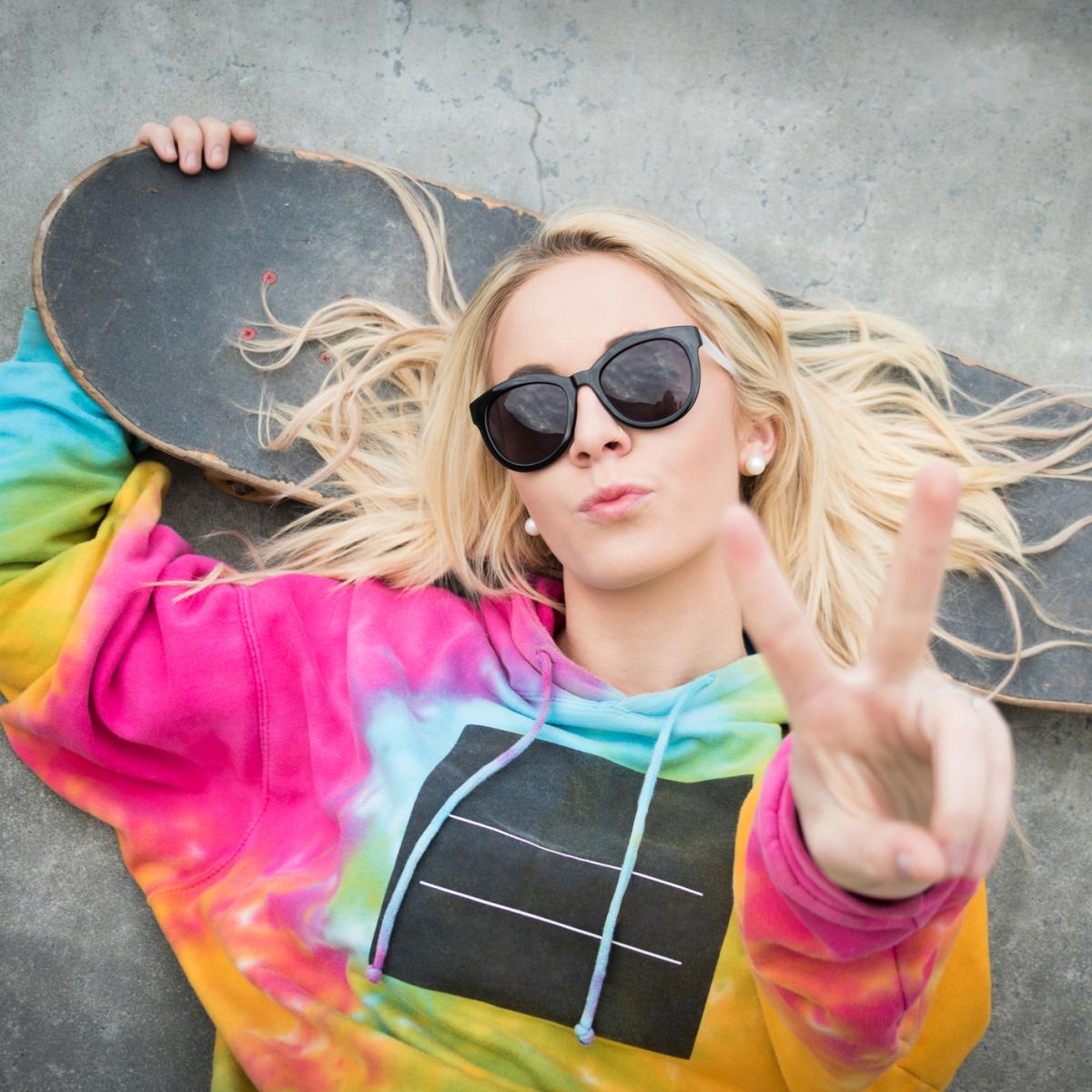 Girl wearing tie dye laying on ground with skateboard and holding a peace sign
